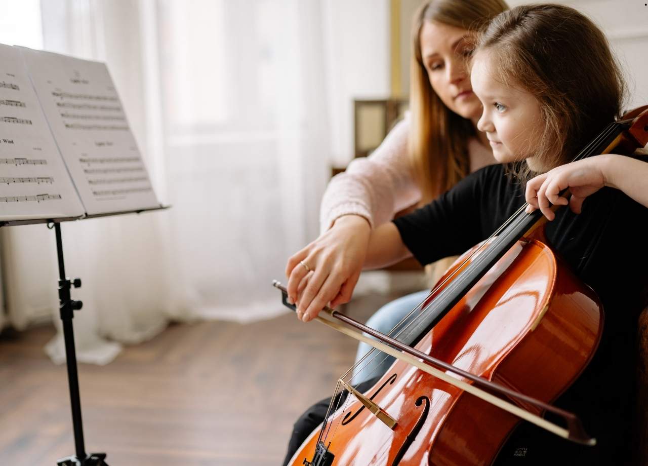 little girl playing chello while looking at music sheet as her mother guides her