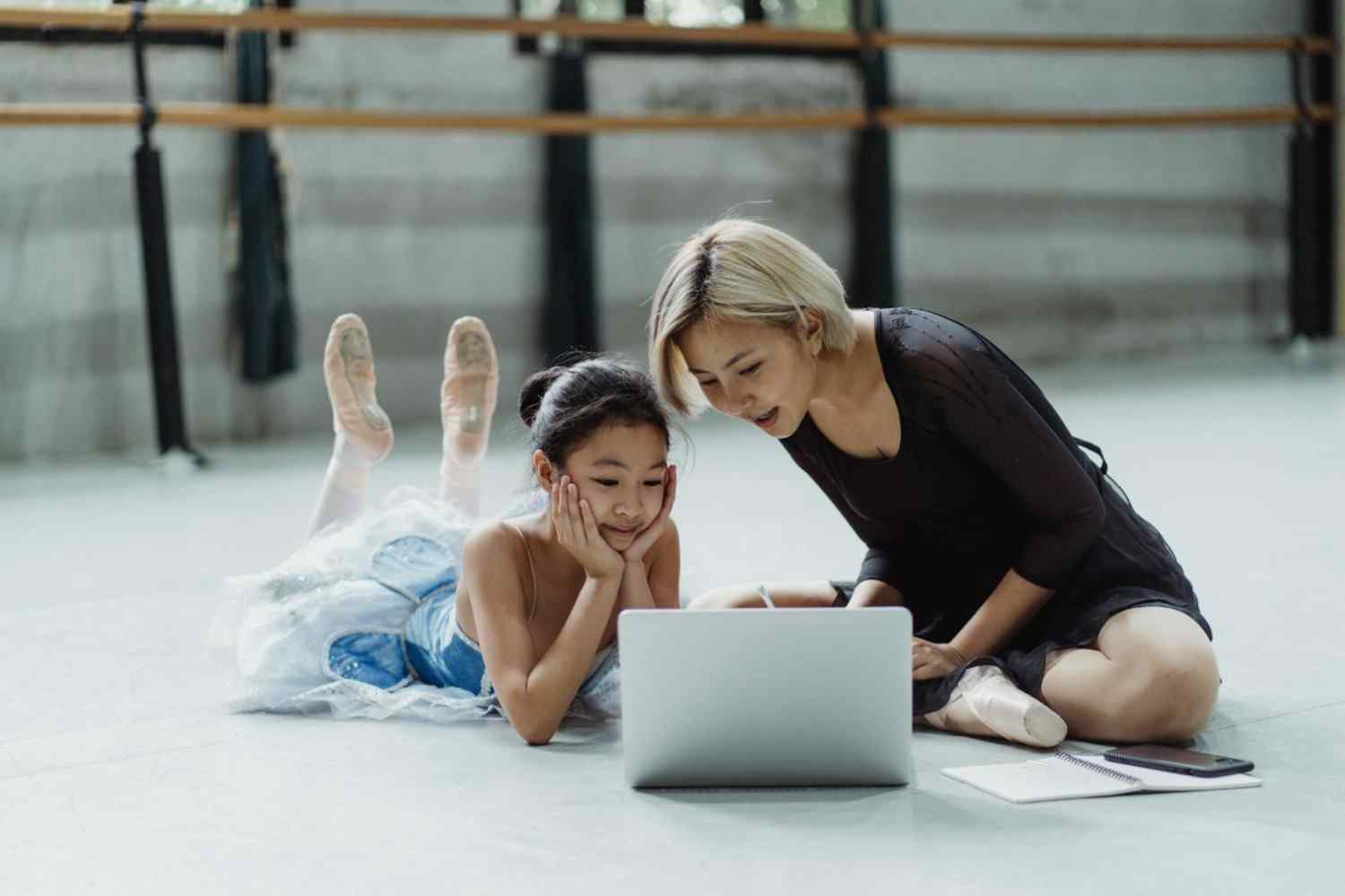 A mother and daughter wearing ballet dance wear as they sit and watch something on a latop