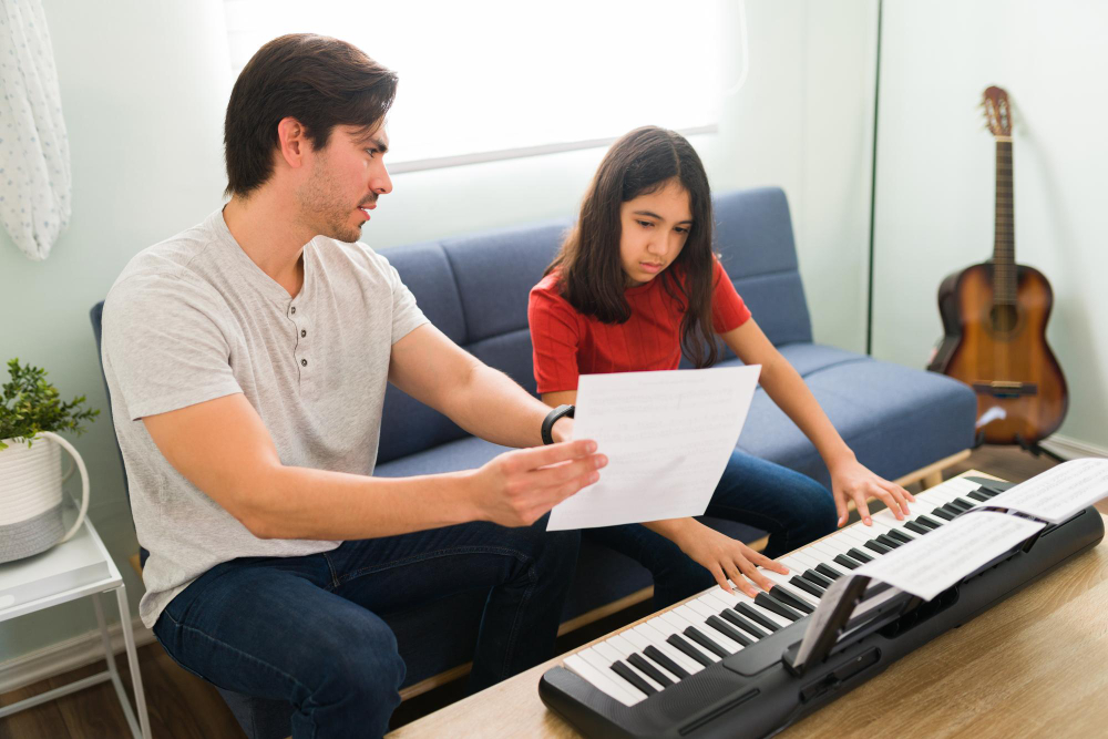 A father holds sheet music for his daughter who is playing a keyboard