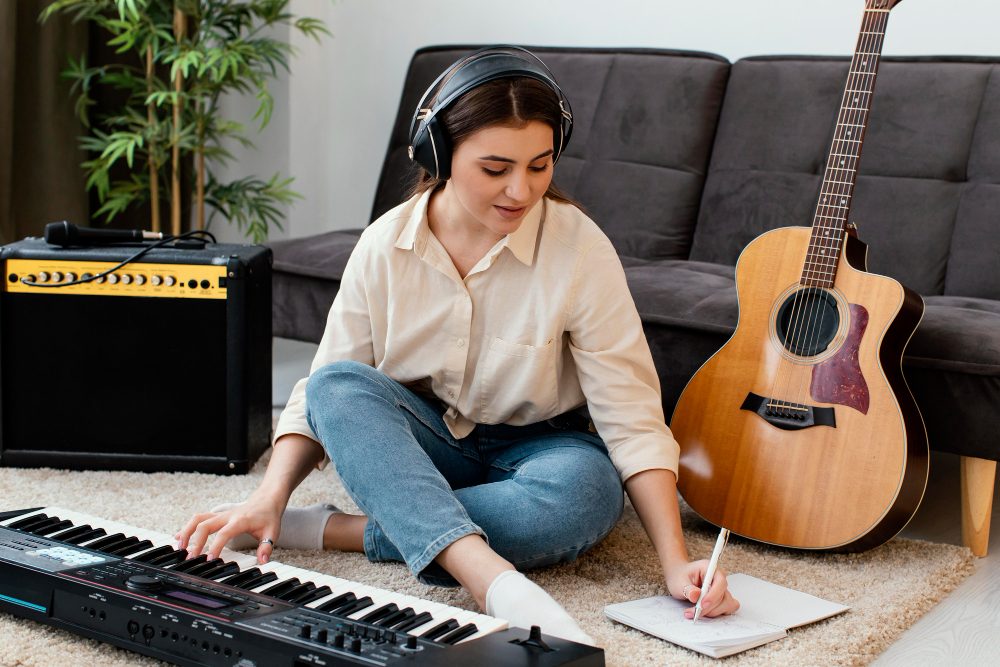 woman sitting on the floor with keyboard, guitar and mic beside her as she jots something in her notebook