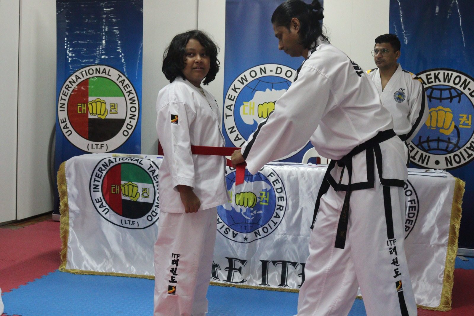 Male student wearing taekwondo uniform with a red belt being tied around his waist by instructor
