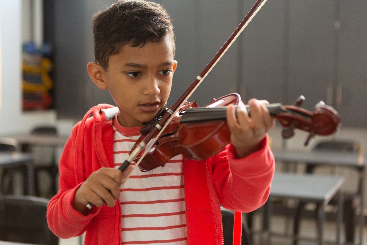 Front view of cute mixed-race schoolboy playing violin in classroom at elementary school