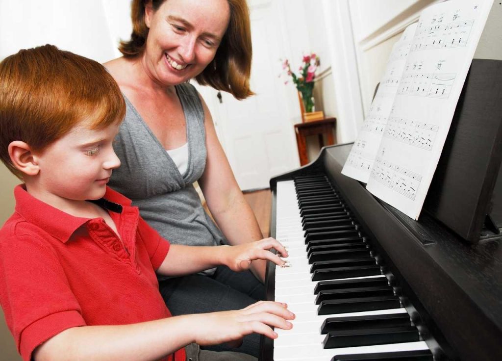 A little boy and his mom smiling as she teaches him how to play piano
