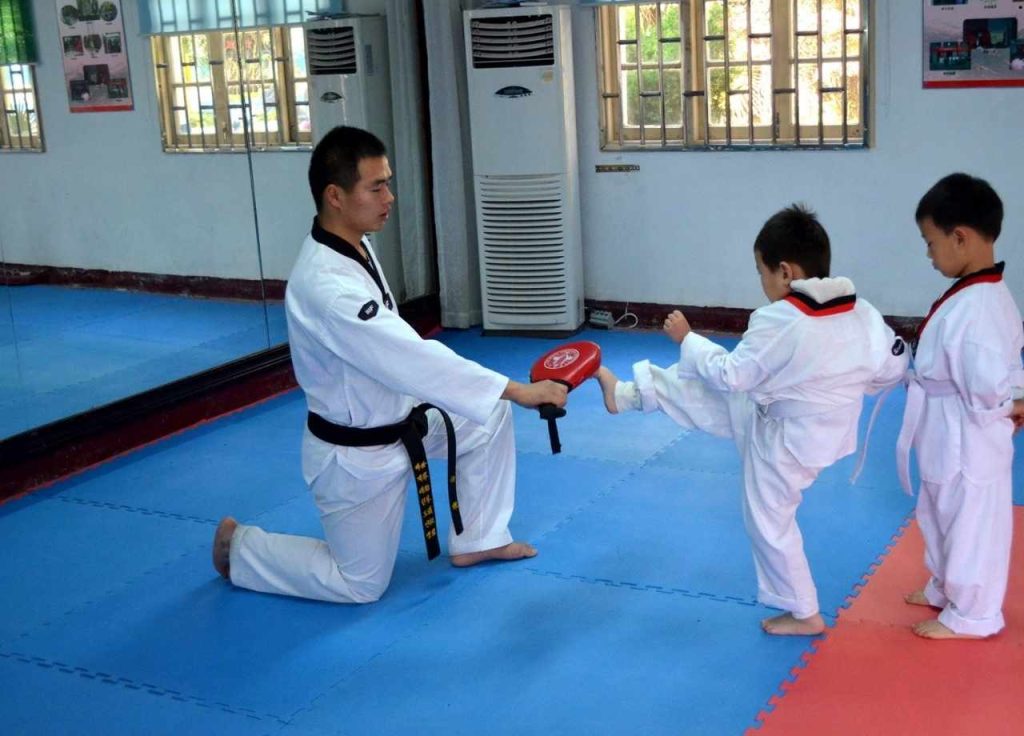 Taekwondo isntructor holding kicking bag as 2 young male students stand in line to practise their kicks